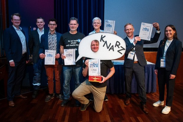 Gruppenfoto der Teilnehmenden des TriboSlam 2025. Die Gruppe steht gemeinsam vor der Kamera; in der Mitte der diesjährige Gewinner Martin Strangfeld, flankiert von den weiteren Vortragenden sowie den Moderatorinnen und Moderatoren.